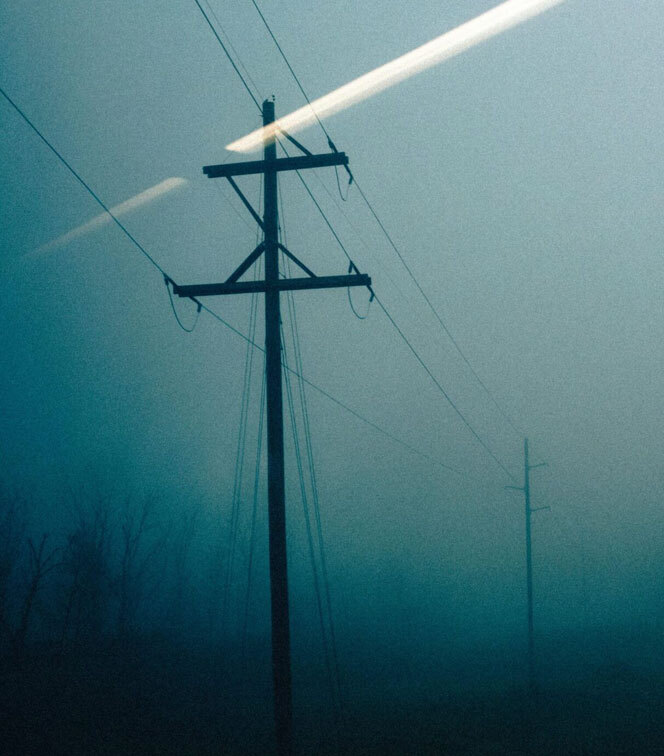 Wooden utility poles with power lines receding into the distance, partially obscured by dense fog under a muted blue sky.
