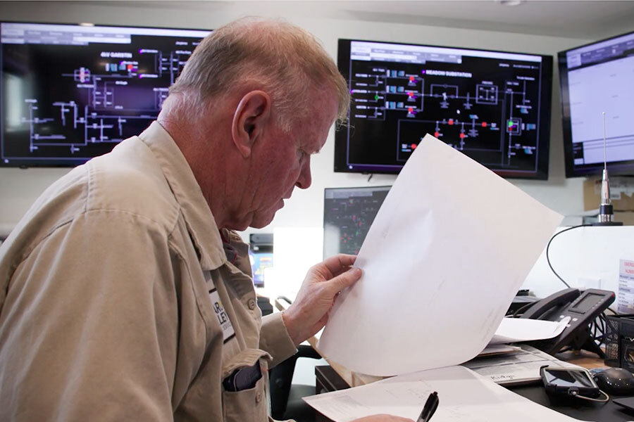 An older man sits at a cluttered desk examining a sheet of paper, with several large monitors behind him displaying complex technical diagrams and system schematics.